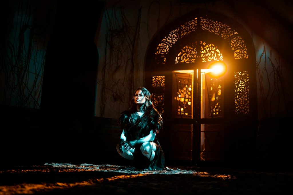 Woman crouching in a dimly lit interior near an ornate arched doorway with warm glowing light.